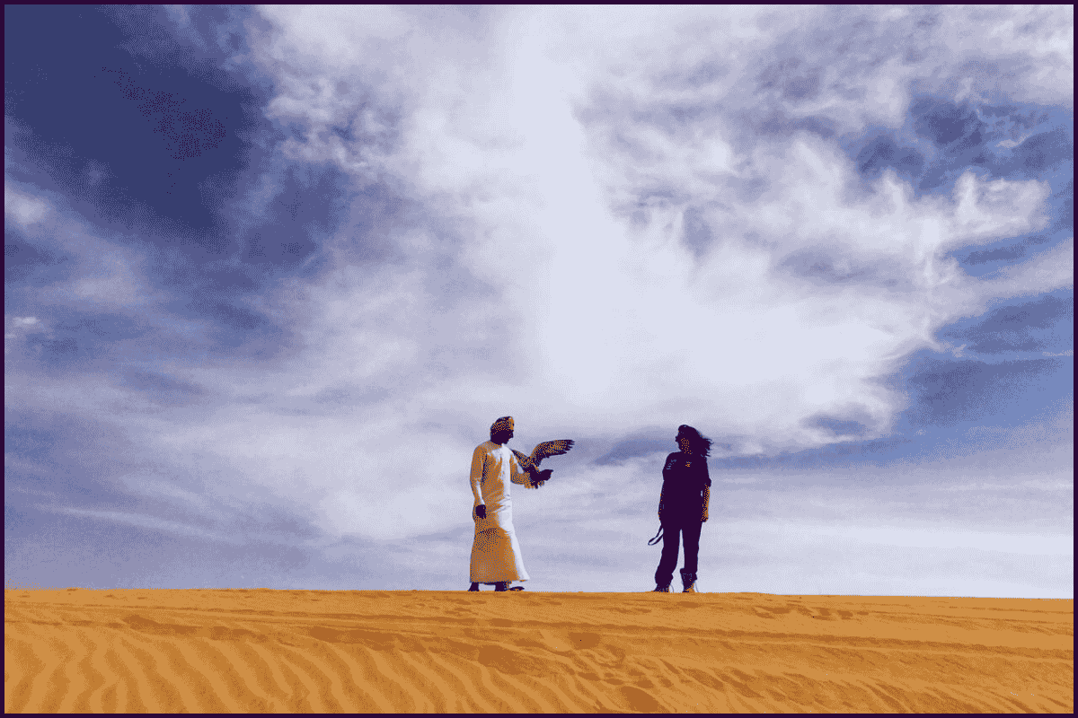 Scottish woman and an Arab falconer stand together on a desert dune under dramatic clouds, symbolising cross-cultural friendship in Arabia.
